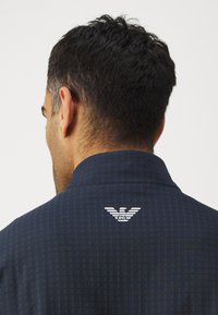 Marineblauwe shirt met een gestructureerd patroon. Bevat een wit logo op de achterkant van de kraag. Materiaal lijkt lichtgewicht met een gestructureerd ontwerp van de kraag.