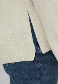 Beiger Strickpullover mit geripptem Saum und seitlichen Schlitzen, kombiniert mit dunkelblauen Jeans aus einem dezent strukturierten Stoff.