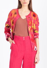 Žena v jasně růžovém květovaném kardiganu, hnědém žebrovaném topu a růžových kalhotách s vysokým pasem a knoflíky, stojící s jednou rukou v kapse.