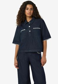 Temno denim srajca s kratkimi rokavi, s belim čipkastim robom na manšetah in žepih, z zapenjanjem na gumbe in sproščenim krojem.