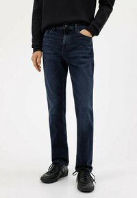 Vaqueros de mezclilla azul oscuro con un corte recto, que cuentan con cinco bolsillos y trabillas para cinturón. Llevados con zapatillas negras, mostrando costuras contrastantes.