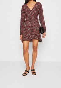 Schwarzes Floral-Wickelkleid mit langen Ärmeln, einem V-Ausschnitt und einem auslaufenden Saum. Kombiniert mit schwarzen, viereckigen, halbhohen Sandalen und einer kleinen Tasche.