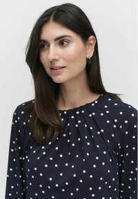 Marineblauwe blouse met witte stippen, voorzien van een geplooide halslijn en lange mouwen, gemaakt van een soepele stof, die een ontspannen pasvorm creëert.