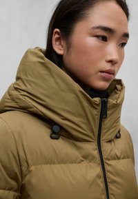 Chaqueta tipo plumón con cuello alto en marrón, que presenta una superficie texturizada, cremallera negra y detalles con cordón. Primer plano de la parte superior.