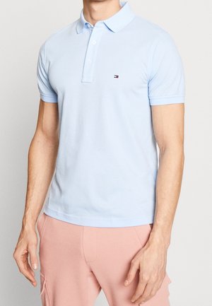 Mann trägt hellblaues Poloshirt mit kurzen Ärmeln und kleinem Logo auf der Brust sowie hellrosa Hose, steht vor einfarbigem Hintergrund.