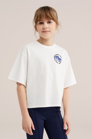 Ung pige med lysebrunt hår iført en hvid, oversized t-shirt med et lille blåt motiv og navy leggings mod en ensfarvet baggrund.