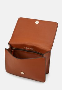 Borsa in pelle marrone con chiusura a pattina, accessori in oro e tasca con cerniera all'interno. Presenta una texture liscia e una stampa logo sottile.