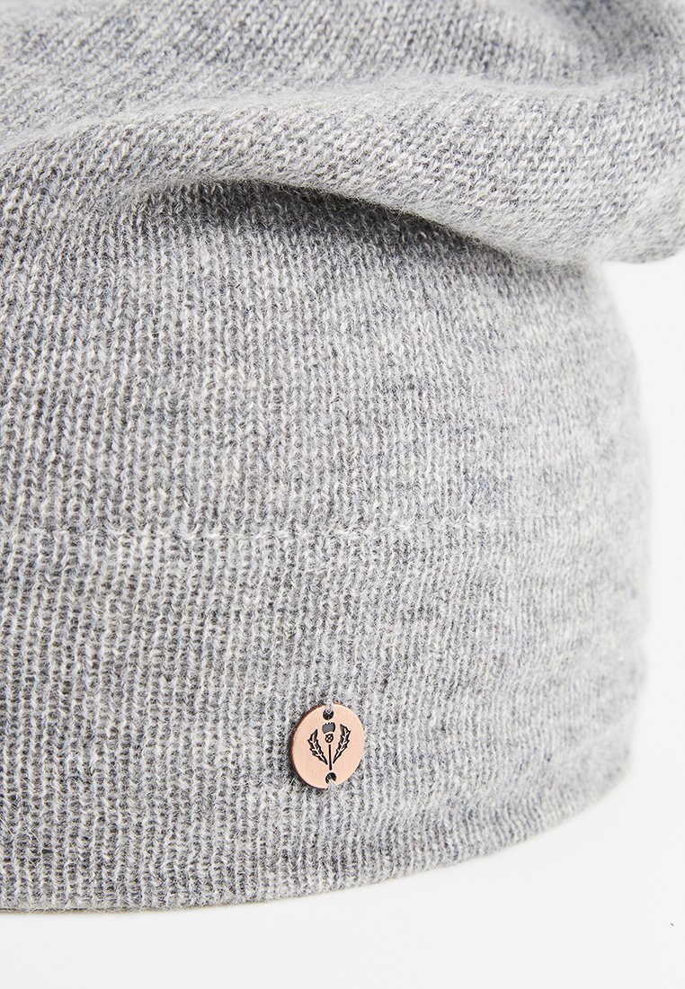 Jasnoszara dziana czapka z małą, okrągłą miedzią metką z stylizowanym wzorem rośliny blisko dolnej krawędzi.