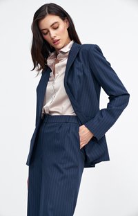 Granatowy garnitur w paski z dopasowaną marynarką i spodniami z wysokim stanem. Pod spodem znajduje się satynowa beżowa bluzka, podkreślająca gładką teksturę.