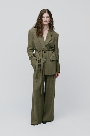 Femme aux cheveux bouclés portant un costume à carreaux vert olive surdimensionné avec un pantalon à jambes larges et un blazer ceinturé, debout devant un fond uni.
