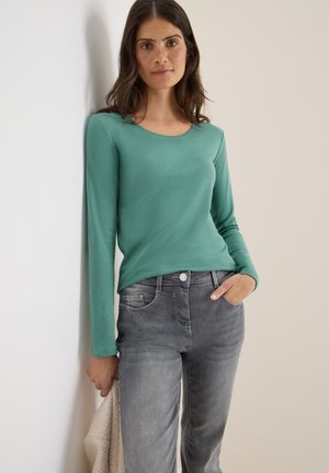 Haut vert à manches longues avec un col rond, associé à un jean gris. Texture de tissu douce, design ajusté et détail de bouton contrastant sur le jean.