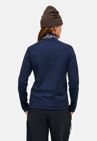 Camicia a maniche lunghe blu navy con colletto alto, dotata di nastro con logo sul collo e maniche raglan. Il materiale appare morbido ed elastico.