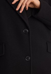 Ręka dostosowująca kołnierz czarnego płaszcza z dwoma dużymi czarnymi guzikami i kieszeniami na klapy, zbliżenie na fakturę tkaniny i przeszycia.