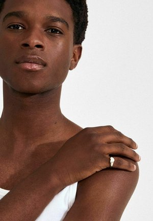 Tuvplāns ar personu, kas vilcienā valkā baltu ribotu tanktopu, rādot brūnu ādu un sudraba gredzenu uz labās rokas, ar gludu, matētu tekstūru.