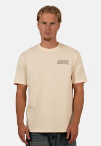 Beige lyhythihainen t-paita, joka on valmistettu puuvillasta ja jossa on pieni "WRONG FRIENDS" -grafiikka vasemmassa rintakohdassa. Klassinen pyöreä kaula-aukko.