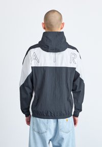 Musta-valkoinen tuulitakkimallinen takki, jossa on takana "AIR"-teksti, korkea kaulus, joustavat hihansuut ja kevyt materiaali.