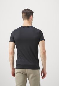 Muž s krátkými tmavými vlasy, oblečený v černé košili s krátkým rukávem a béžových kalhotách, stojící čelem ke bílé zdi s malým tetováním na paži.