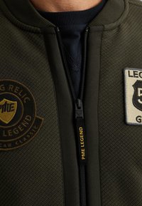 Veste bomber texturée verte avec un col en tricot noir, fermeture éclair avec un tirette étiquetée "PME LEGEND" et deux patchs circulaires.