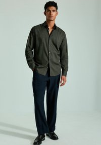 Dunkelgrünes Hemd mit Knöpfen und geometrischem Muster, kombiniert mit navyblauen Hosen und schwarzen Schuhen. Glattes Gewebe, taillierter Schnitt.