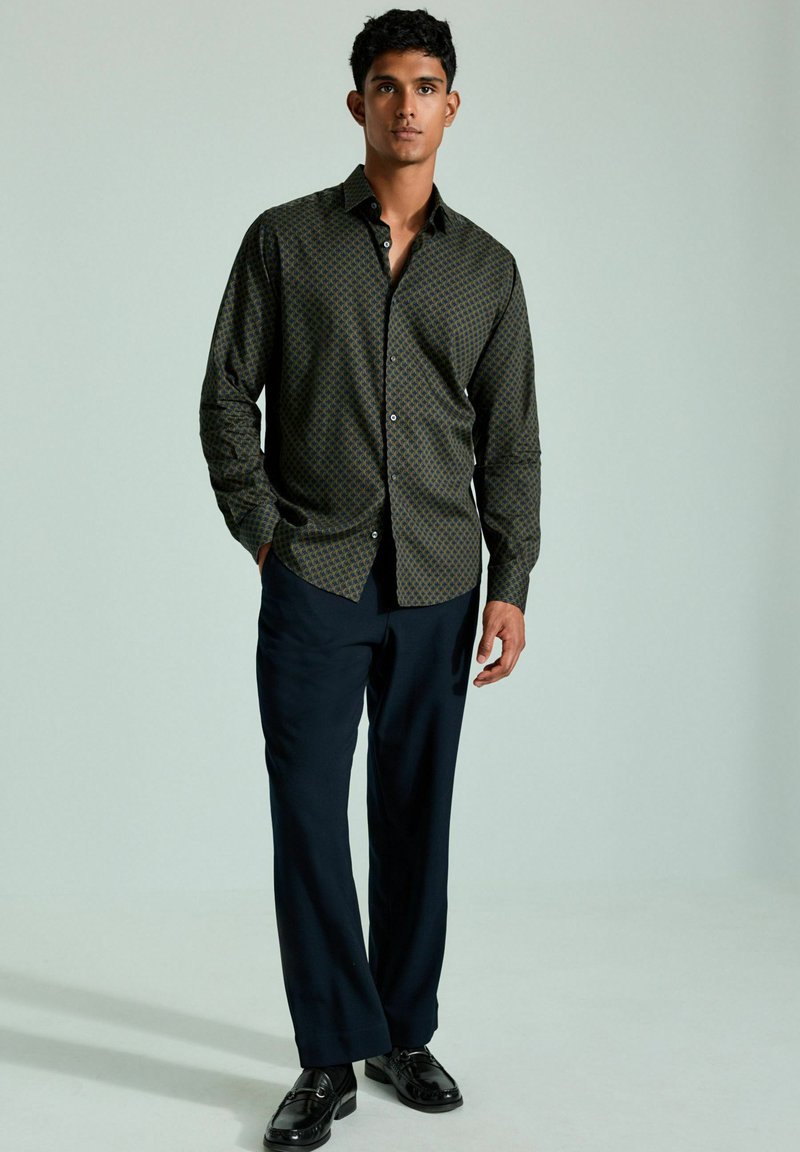 Dunkelgrünes Hemd mit Knöpfen und geometrischem Muster, kombiniert mit navyblauen Hosen und schwarzen Schuhen. Glattes Gewebe, taillierter Schnitt.