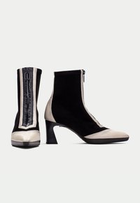 Svarta mockaskor med beige lackläderdetaljer, dragkedjestängning och medelhög klack, med en texturerad svart randdetalj.