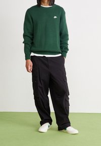 Person, der einen grünen Nike-Pullover, schwarze Cargo-Hosen und weiße Turnschuhe trägt, steht auf einem grünen Boden vor einer weißen Wand.