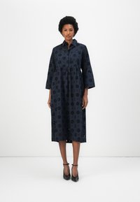 Abito in cotone blu navy con maniche 3/4, motivo floreale ricamato, colletto e orlo al ginocchio. Indossato con scarpe con tacco alto nere.