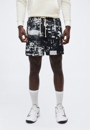 Mand iført sorte og hvide atletikshorts med abstrakt mønster, hvide crew-sokker, hvide sneakers og en cremefarvet sweatshirt.