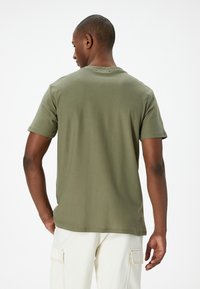 Camiseta de algodón verde oliva con mangas cortas y cuello redondo, con un corte relajado y una textura suave. Detalles de costura visibles.