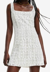 Weißes Spitzen-Minikleid mit quadratischem Ausschnitt, ärmellosem Design, filigranen Blumenmustern und leicht ausgestellter A-Linie.