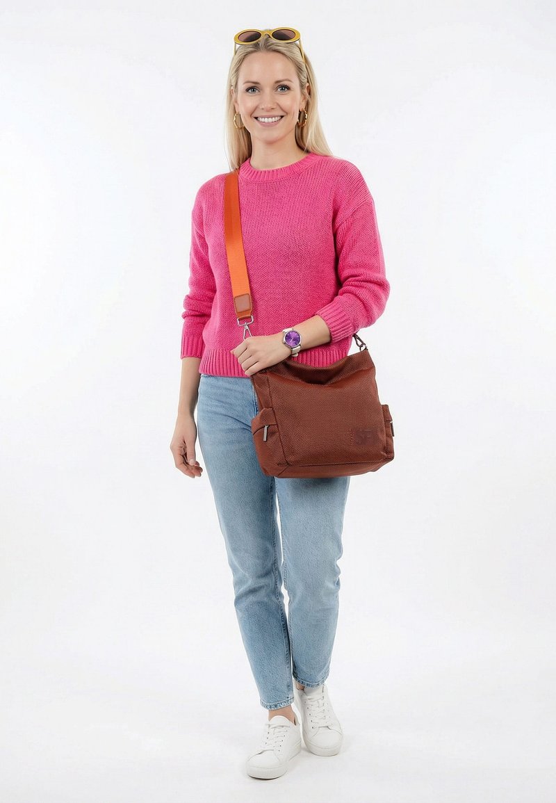 Mujer sonriente con cabello rubio que lleva un suéter rosa, jeans azul claro, zapatillas blancas y un bolso cruzado marrón con correa naranja.