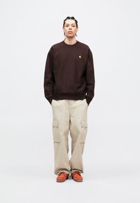 Felpa marrone scuro con un piccolo logo, abbinata a pantaloni cargo beige chiaro con molteplici tasche e scarpe arancioni con lacci.