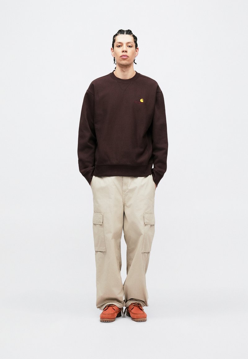 Dunkelbrauner Pullover mit kleinem Logo, kombiniert mit hellbeigen Cargohosen mit mehreren Taschen und orangefarbenen Schnürschuhen.