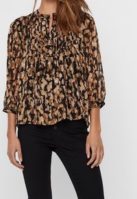Blouse noire avec un motif floral dans des tons de marron et d'or. Présente un design plissé, des manches 3/4 et un décolleté en forme de clé. Accompagnée d'un pantalon noir.