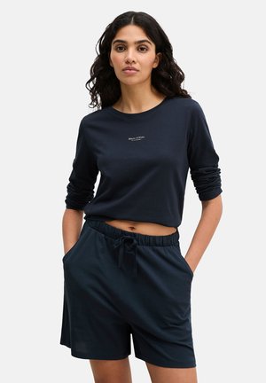 Jeune femme aux cheveux bouclés et foncés, portant un t-shirt manches longues bleu marine et un short assorti, mains dans les poches, devant un fond uni.