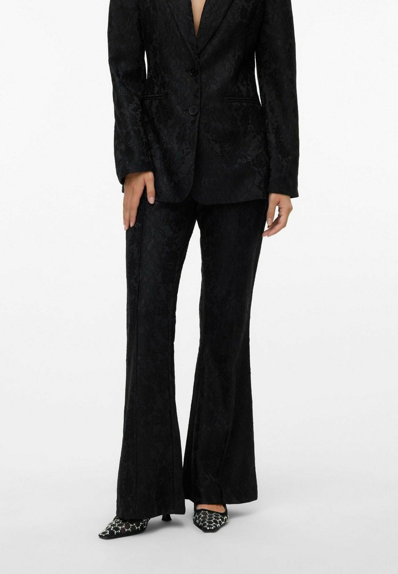 Costume noir avec texture florale, comprenant un blazer à un bouton et un pantalon évasé. Assorti à des chaussures à motif noir et blanc à bout pointu.
