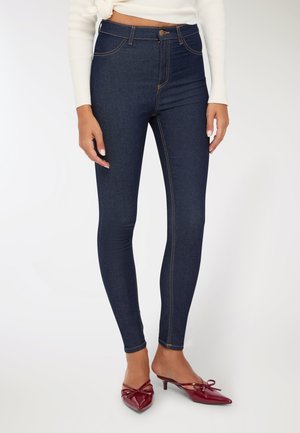 Donna che indossa jeans skinny blu scuro, maglietta bianca a maniche lunghe e scarpe con tacco a punta rosse, in piedi su sfondo bianco.