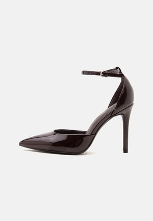 Chaussure à talon haut marron foncé brillante avec bout pointu et bride de cheville avec boucle argentée sur fond blanc uni.