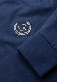 Camiseta azul marino de manga larga con letras blancas bordadas "EX" dentro de una corona de laurel en la zona del pecho.