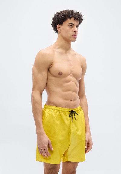 Νεαρός άνδρας με σγουρά μαλλιά που φοράει κίτρινα μαγιό με μαύρο κορδόνι, στέκεται μπροστά σε ένα απλό φωτεινό φόντο, κοιτώντας προς τα δεξιά.