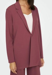 Donna in blazer oversize color rosa polveroso sopra una maglietta bianca, con pantaloni larghi abbinati, in piedi contro uno sfondo neutro.