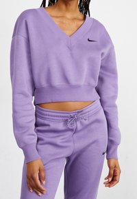 Kvinna iförd en lavendelfärgad V-ringad kort sweatshirt och matchande sweatpants med snörning och svarta Nike-logotyper mot en vit bakgrund.