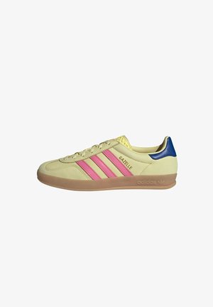 Béžové tenisky Adidas Gazelle se třemi růžovými pruhy. Mají modrý akcent na patě a gumovou podrážku. Hladký semišový svršek.