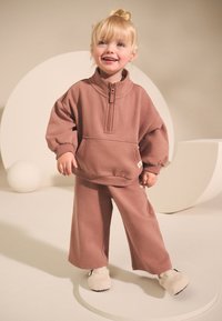 Felpa in pile marrone chiaro con collo alto, cerniera e tasca frontale abbinata a pantaloni ampi coordinati. Il modello indossa scarpe bianche e pelose.