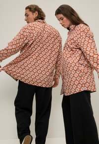 Camisas de manga larga en color óxido con patrón de diamantes blancos, corte relajado. Combinadas con pantalones negros. Ambas camisas tienen un cuello abotonado y un dobladillo curvo.