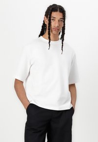 T-shirt en coton blanc à manches courtes, avec un col rond, coupe décontractée et texture subtile. Associé à un pantalon noir.