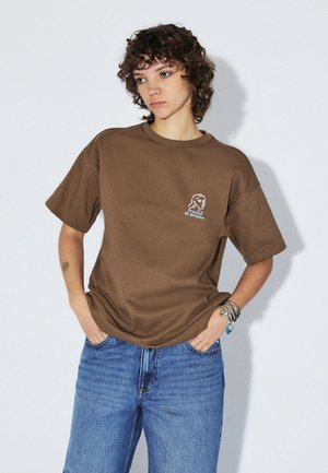 Młoda osoba z kręconymi włosami, ubrana w brązowy luźny t-shirt z białym logo i niebieskie dżinsy, ręce w kieszeniach, na jednolitym tle.