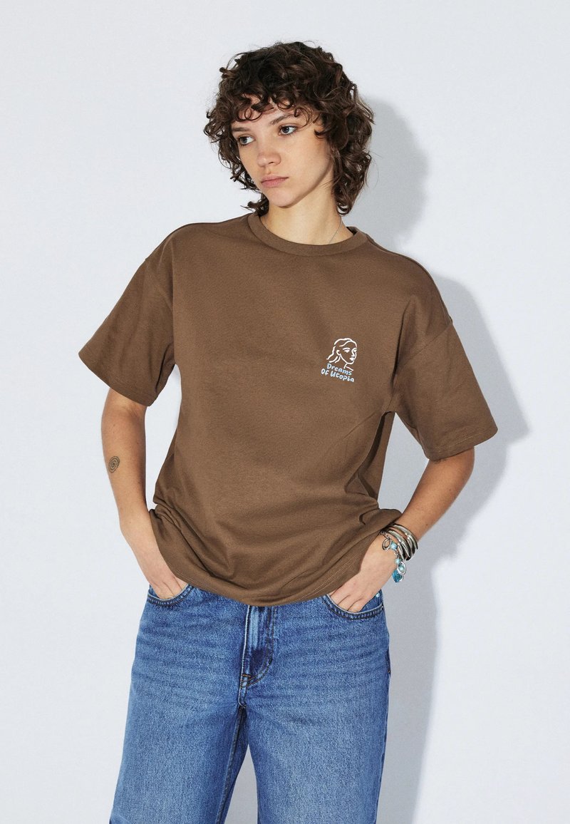 Młoda osoba z kręconymi włosami, ubrana w brązowy luźny t-shirt z białym logo i niebieskie dżinsy, ręce w kieszeniach, na jednolitym tle.