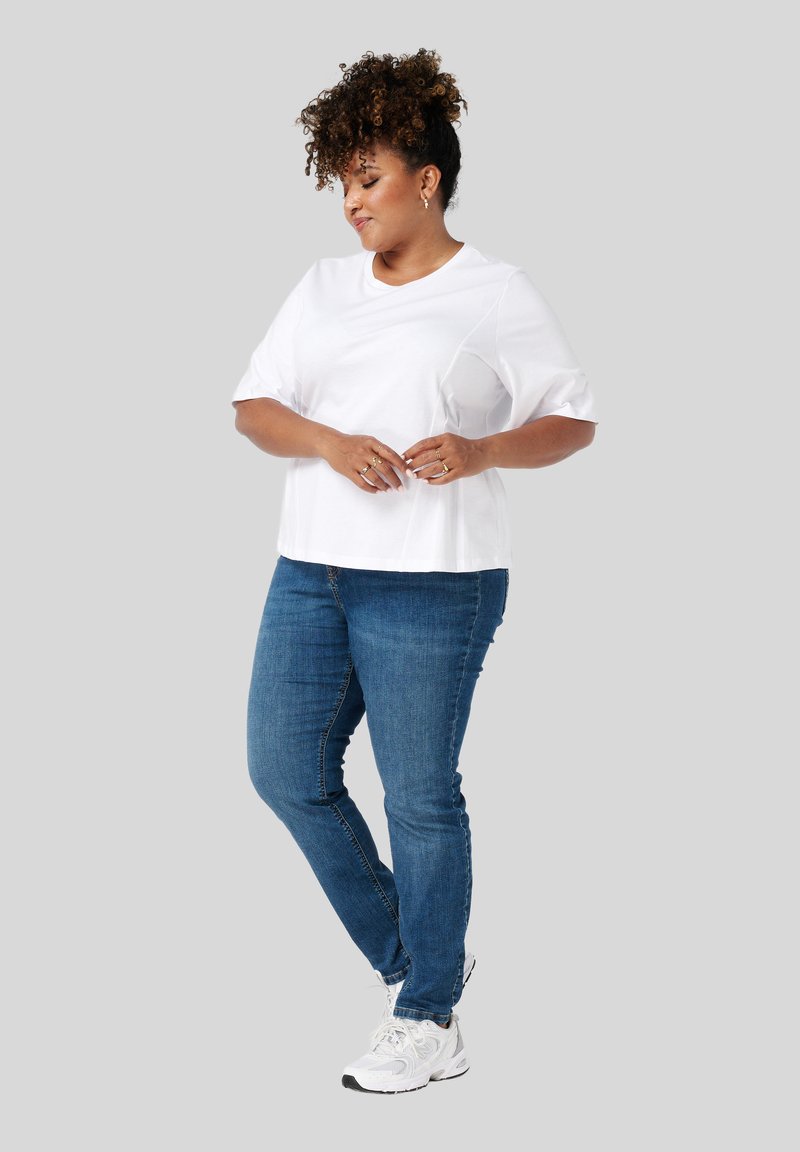 Vrouw met krullend haar, gekleed in een witte shirt met korte mouwen, blauwe jeans en witte sneakers, staat met gekruiste handen, kijkt omlaag, op een grijze achtergrond.