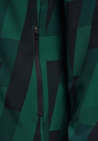 Ritsluiting in het zwart op een donkergroen en zwart patroon stof met geometrische vormen en een gladde textuur.